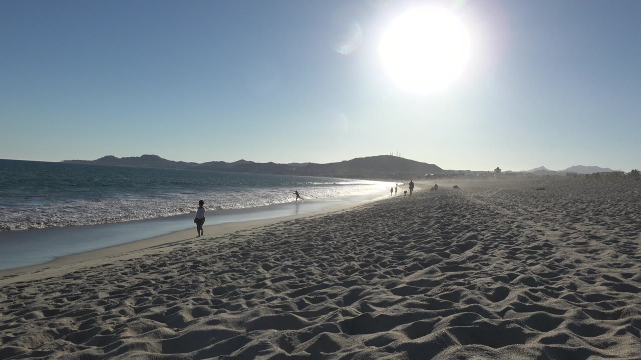Los Cabos shoreline view with one person standing or walking near the wet sand, waves breaking at left, sun reflected on the water, and several distant beachgoers farther down the beach.