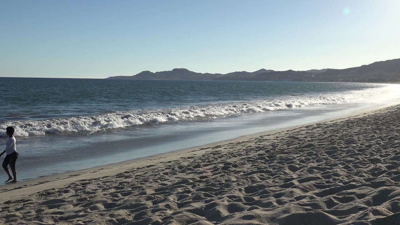 A person walks near the left edge of a Los Cabos beach frame, with rippled sand, white surf, blue sea, distant hills, and bright sun glare on the water.