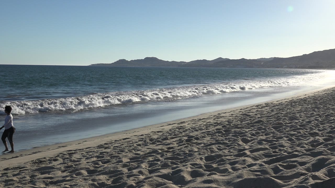 A person walks through the lower middle of the Los Cabos beach view while white waves roll behind them and sunlight reflects off the sea.