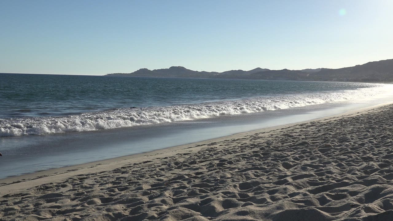 Empty sandy beach and surf with distant hills across the water.