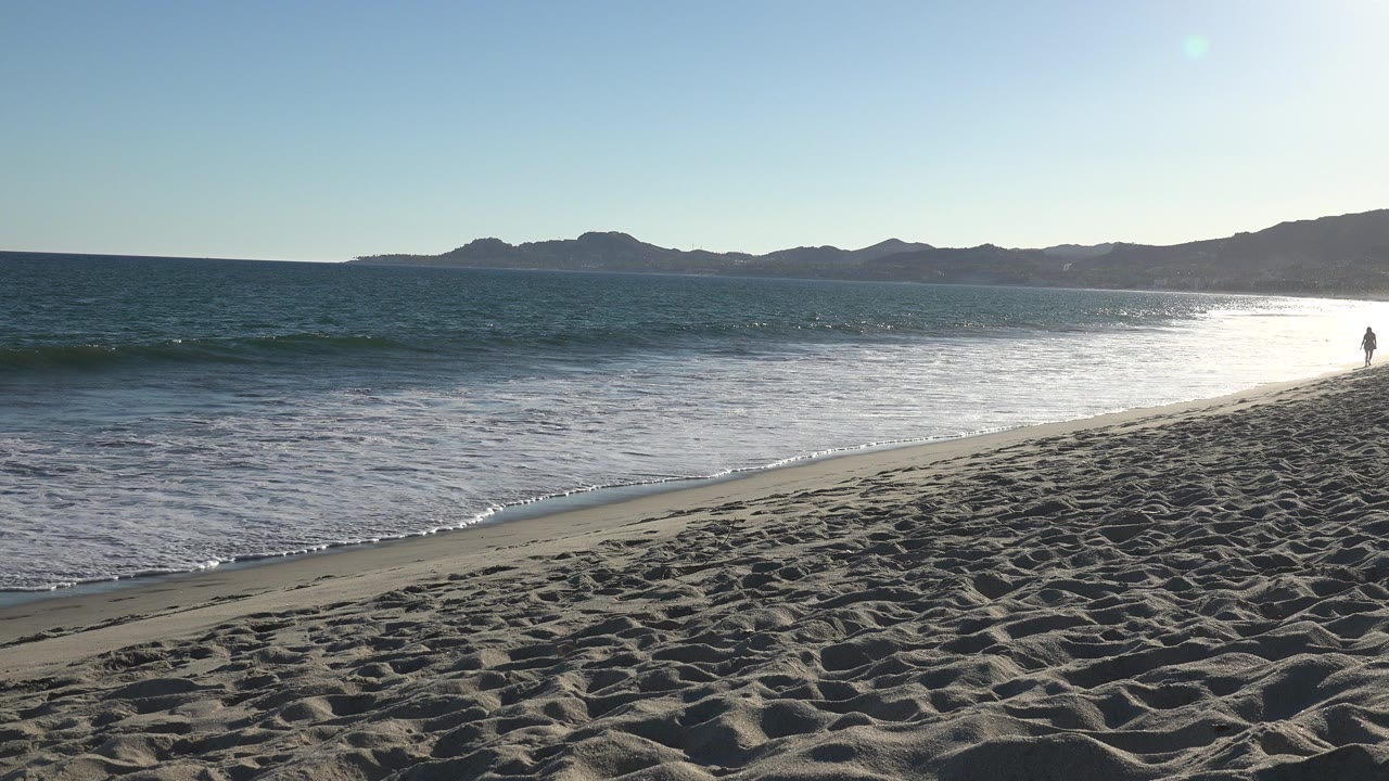 Wide beach with small waves, bright water glare, and mountains on the horizon.