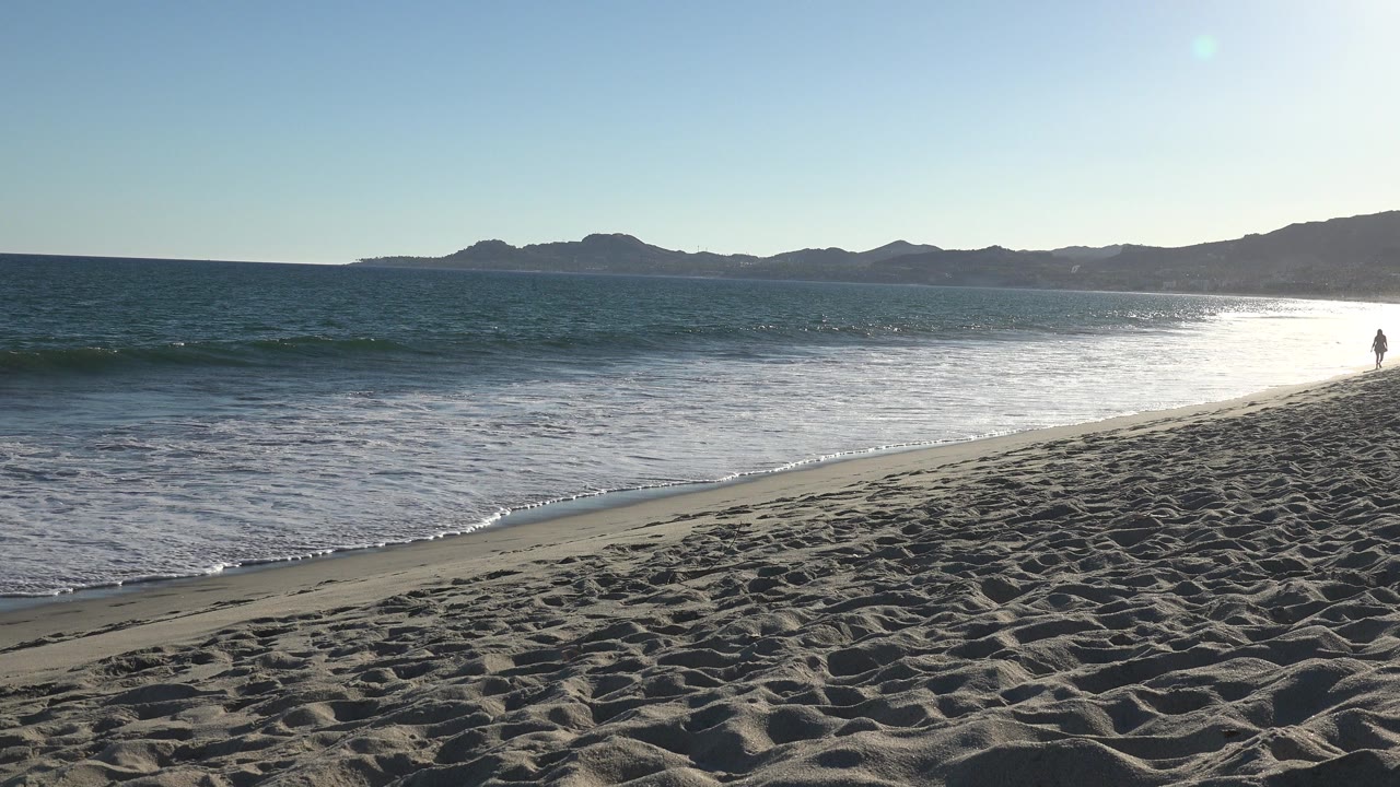 Los Cabos shoreline with low waves nearing the wet sand, dry textured sand in front, a dark mountain ridge across the horizon, and one tiny distant figure near the right edge.