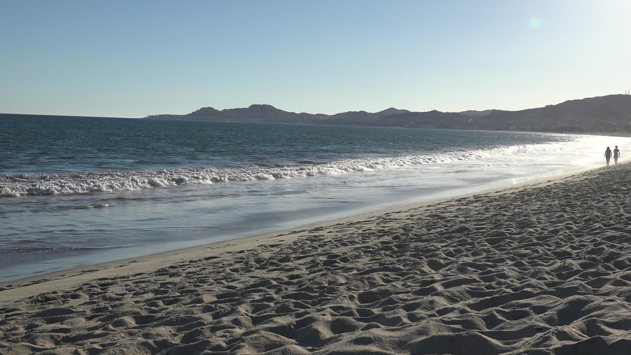 Opening view of a Los Cabos beach with rippled sand in the foreground, white surf at low tide, distant hills across the water, and two small people walking near the right shoreline.