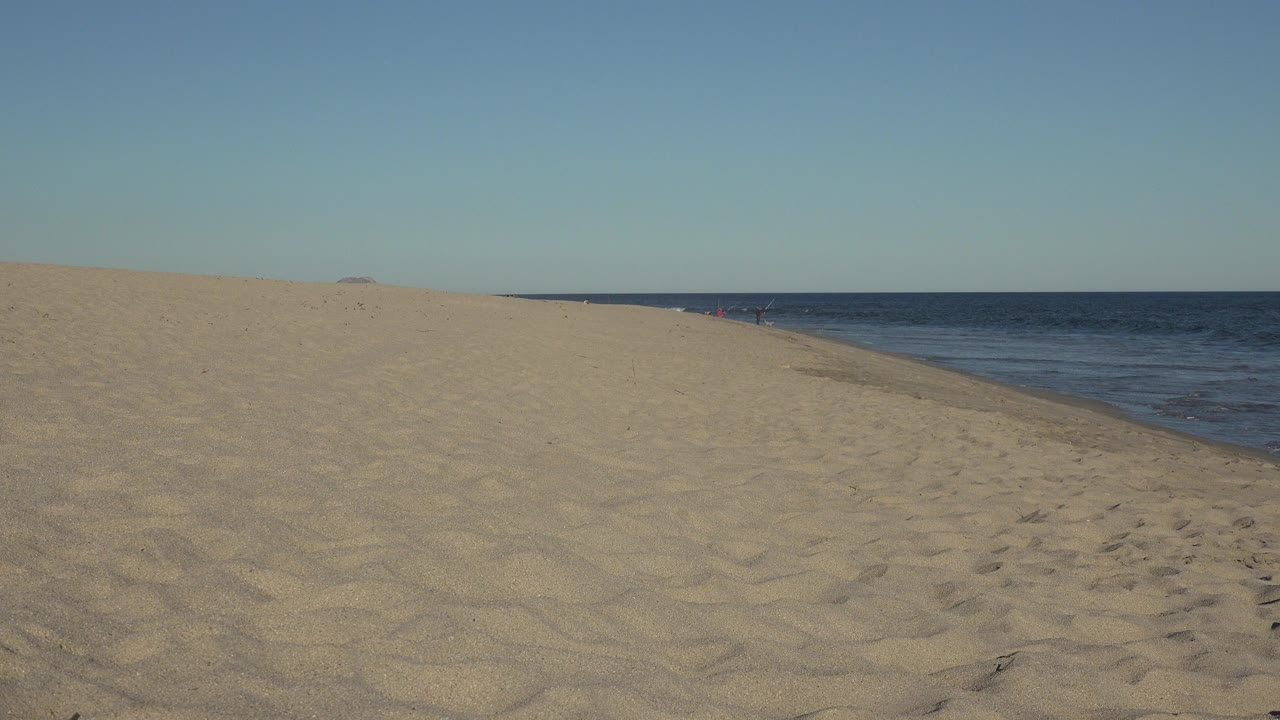 Opening view of a Los Cabos sandy beach with the ocean running along the right side, pale blue sky above, and no visible people.