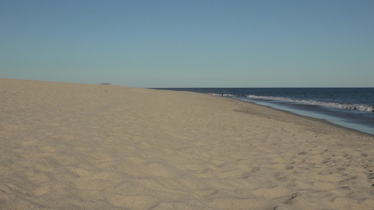 Opening frame of a wide Los Cabos beach view with rippled sand in front, waves along the right side, blue ocean on the horizon, and no visible people.