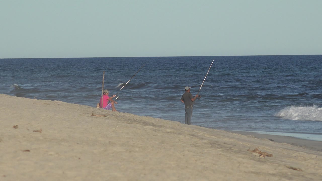 The standing angler holds a rod toward the water while another person sits farther left on the Los Cabos sand near a second upright rod.