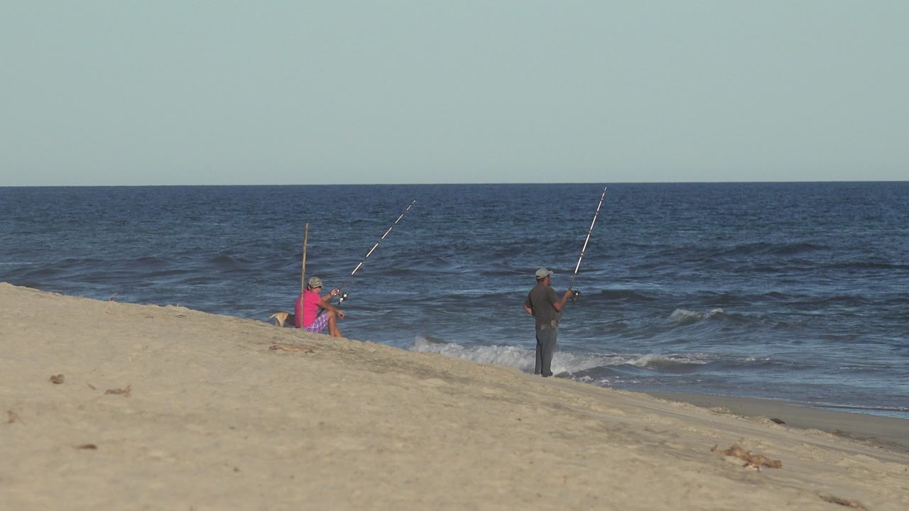 Two people fishing from a sandy beach beside ocean waves.