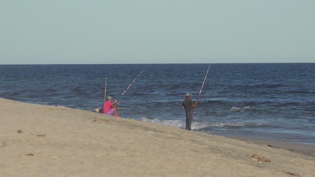 One second in, the two fishers remain on the Los Cabos sand, with the person in bright red or pink near gear and the person in dark clothing closer to the waterline.