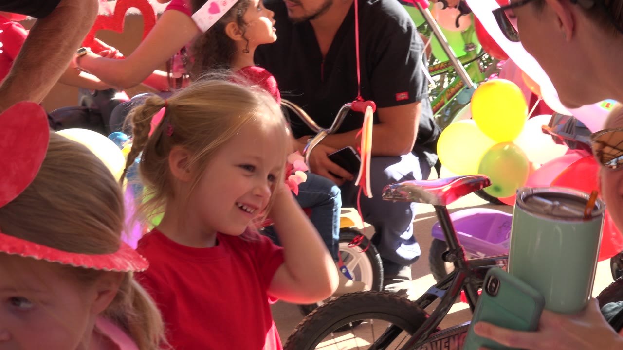 A smiling young girl in a red shirt is framed close to camera in Los Cabos, Mexico, with adults, balloons, and a children's bicycle visible behind her.