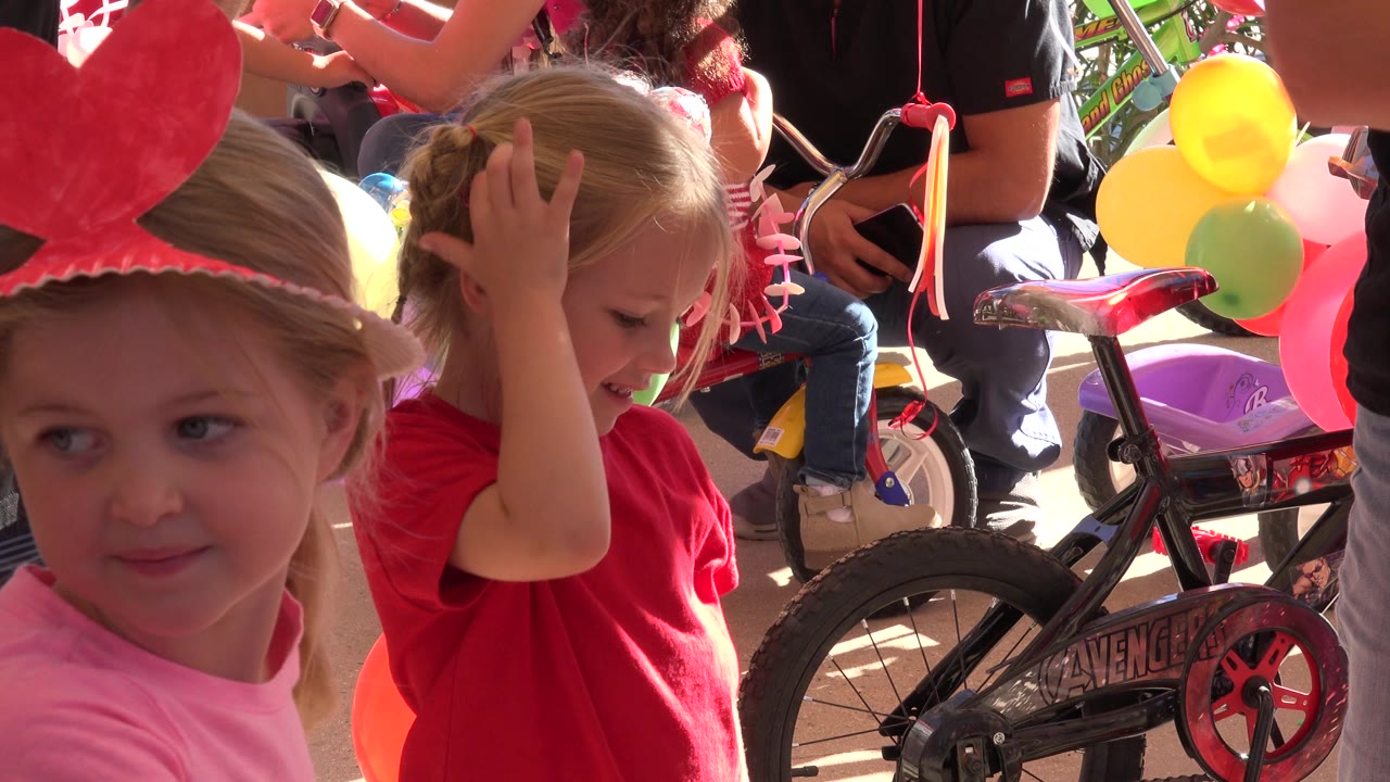 At 0:00 in Los Cabos, Mexico, a young girl in a pink shirt and red visor stands in the foreground while a blonde girl in a red shirt touches her head near balloons and a children's bicycle.