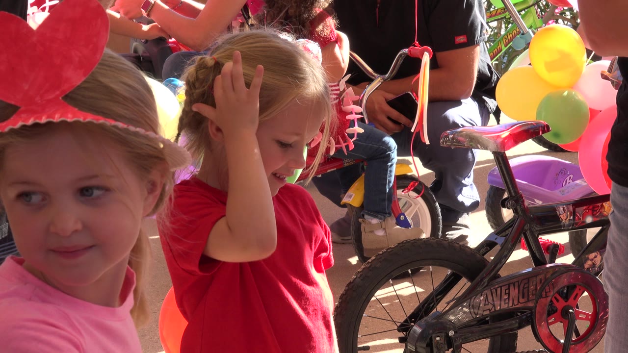 At 0:01 in Los Cabos, Mexico, the girl in the red shirt keeps her hand on her head while the girl in pink remains partly visible beside bright balloons and the black children's bicycle.