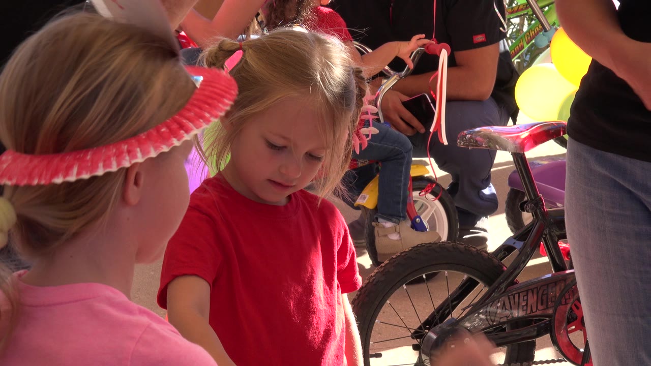 In Los Cabos, Mexico, two young girls stand close together near a small bicycle, balloons, and adults during a festive preparation moment.
