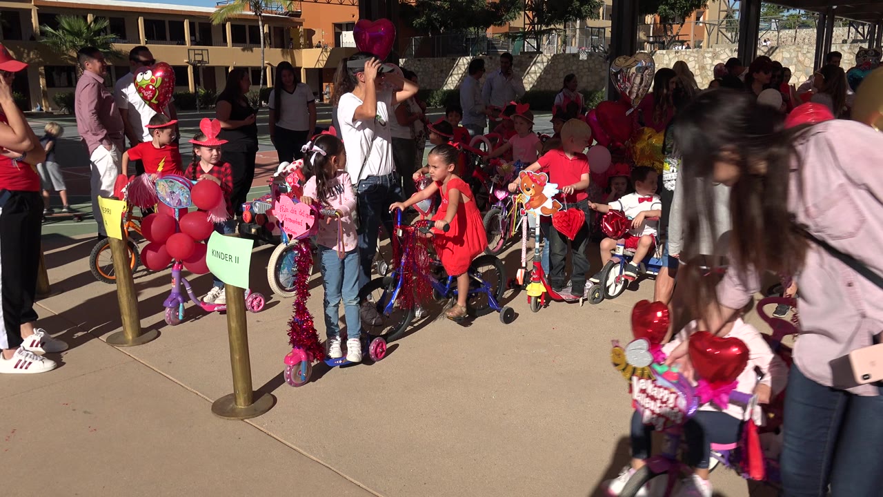 A woman in a pale top leans into the foreground while children, adults, balloons, and decorated bikes fill the sunny Los Cabos path behind her.
