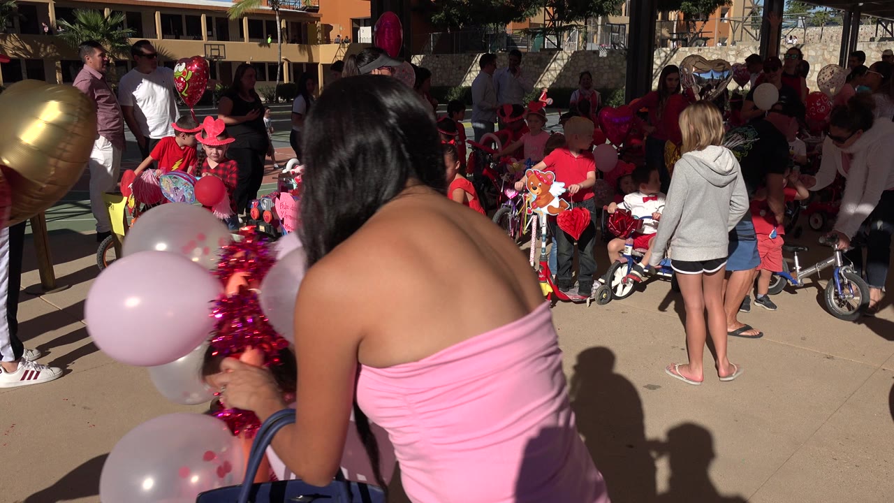 Children and parents at the Los Cabos party cluster around red and pink balloons, small bicycles, and party decorations while a woman remains close in the foreground.