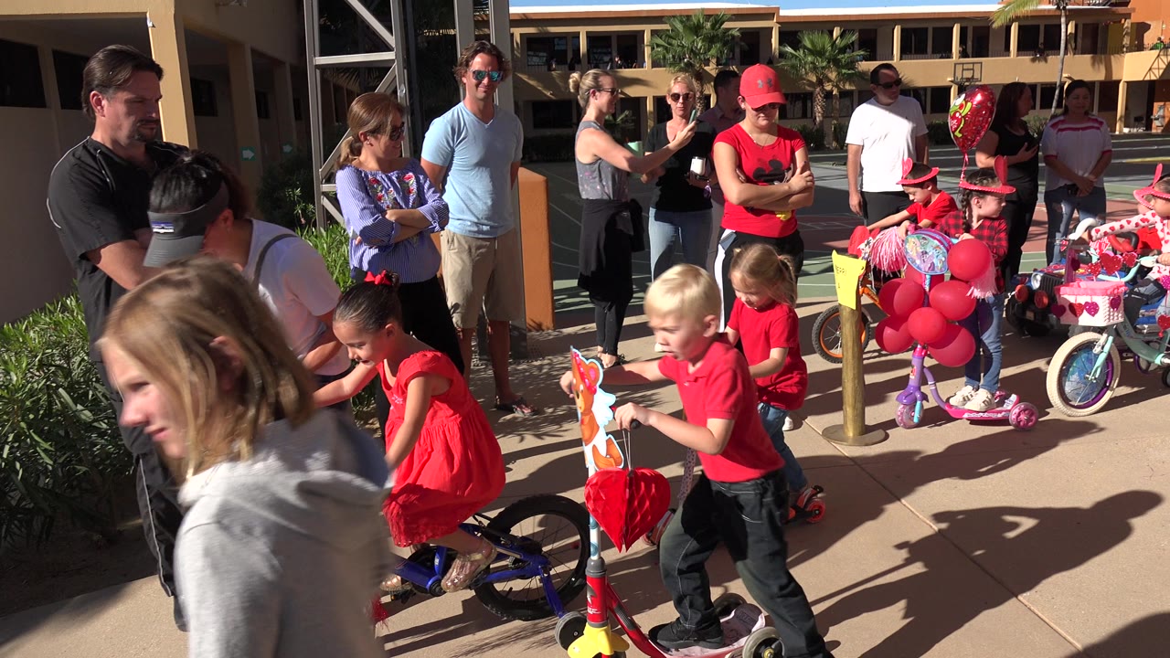 Children continue riding decorated scooters and bicycles in Los Cabos, Mexico, with a red balloon cluster, heart ornaments, adults, shrubs, and a building exterior behind them.