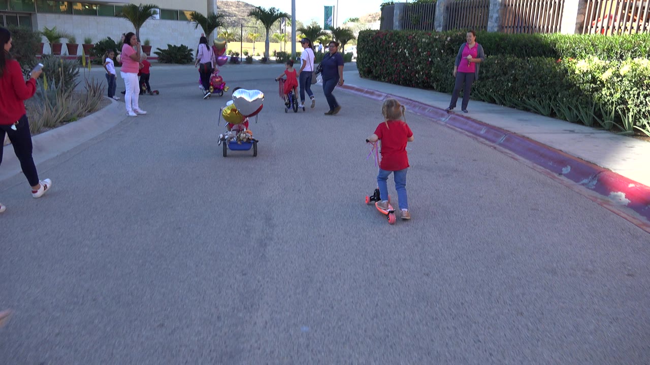 Child in red shirt on a scooter facing a group of adults and children outside a building.