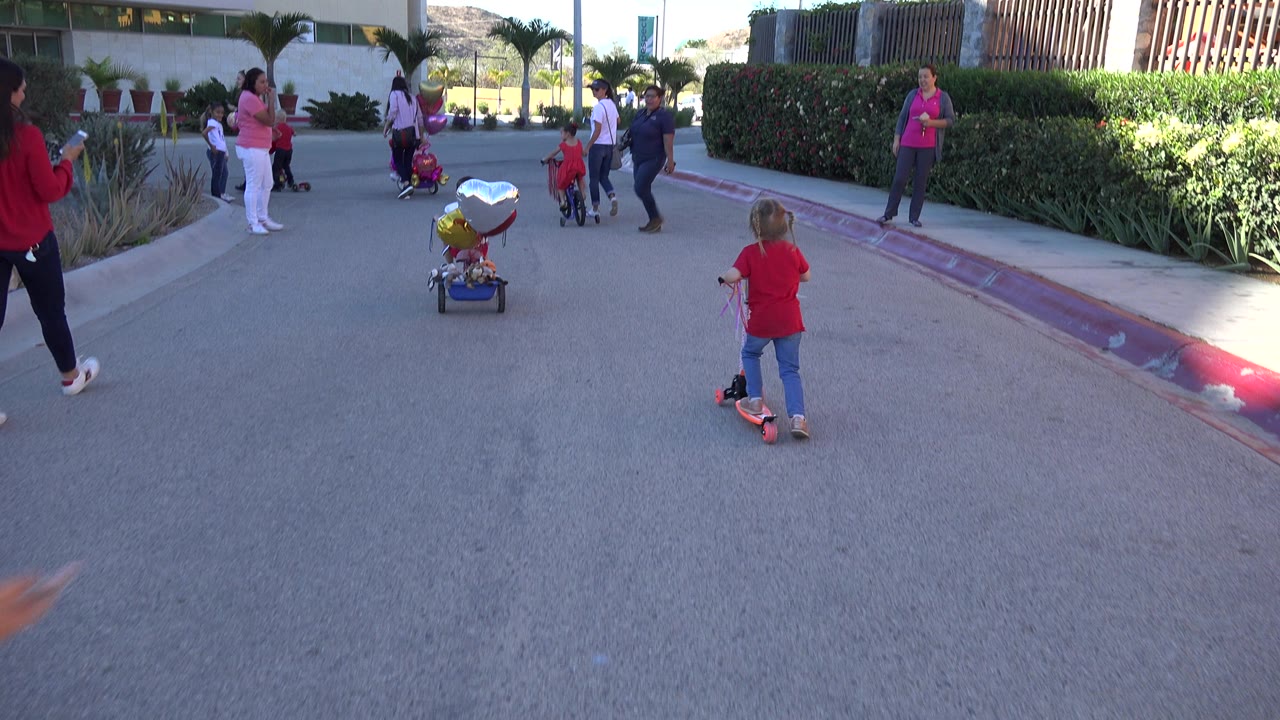 In Los Cabos, Mexico, the child in red stays centered on the scooter while a small group with decorated ride-on toys and balloons stands outside the signed building.