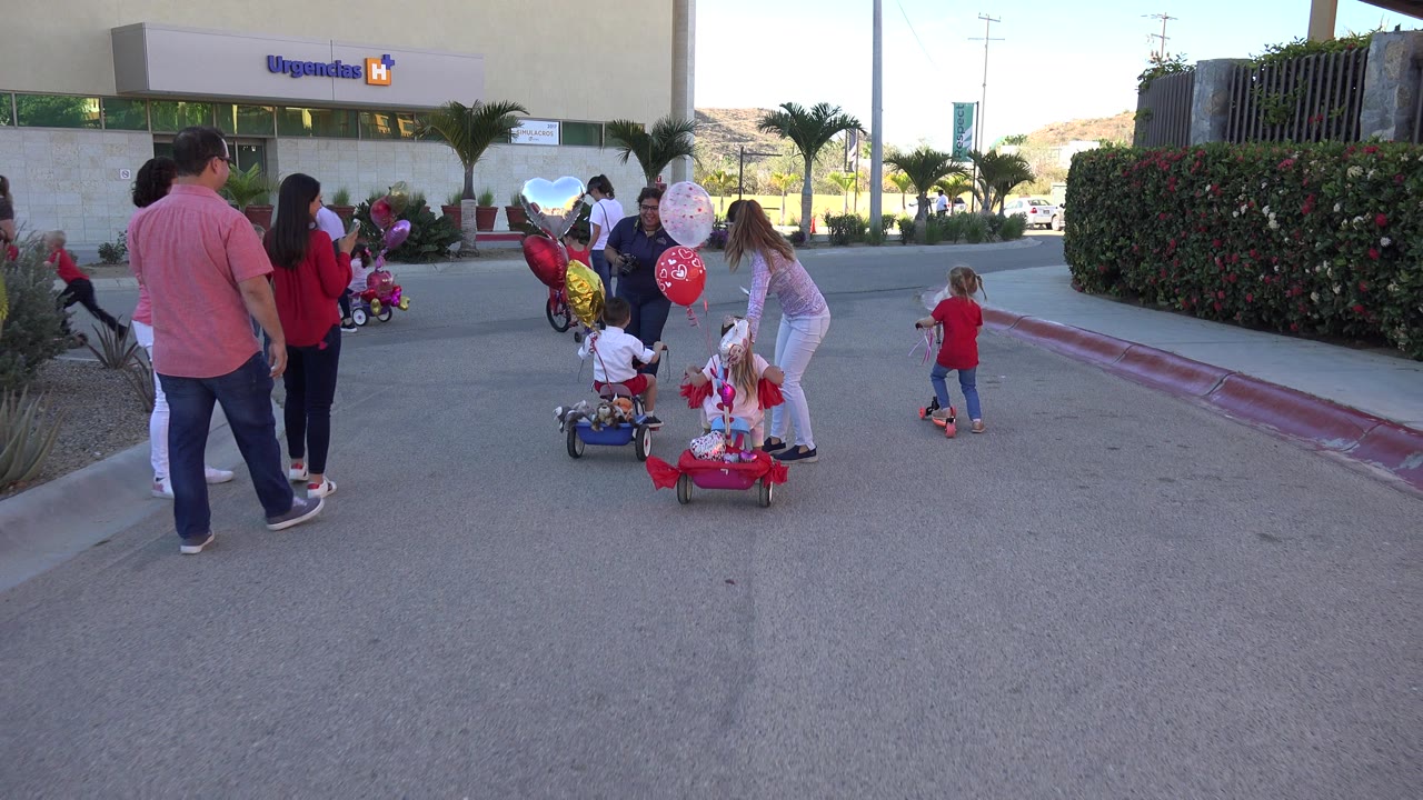 In Los Cabos, Mexico, two children sit on small decorated bikes with balloons while adults stand nearby outside a building entrance marked Urgencias.
