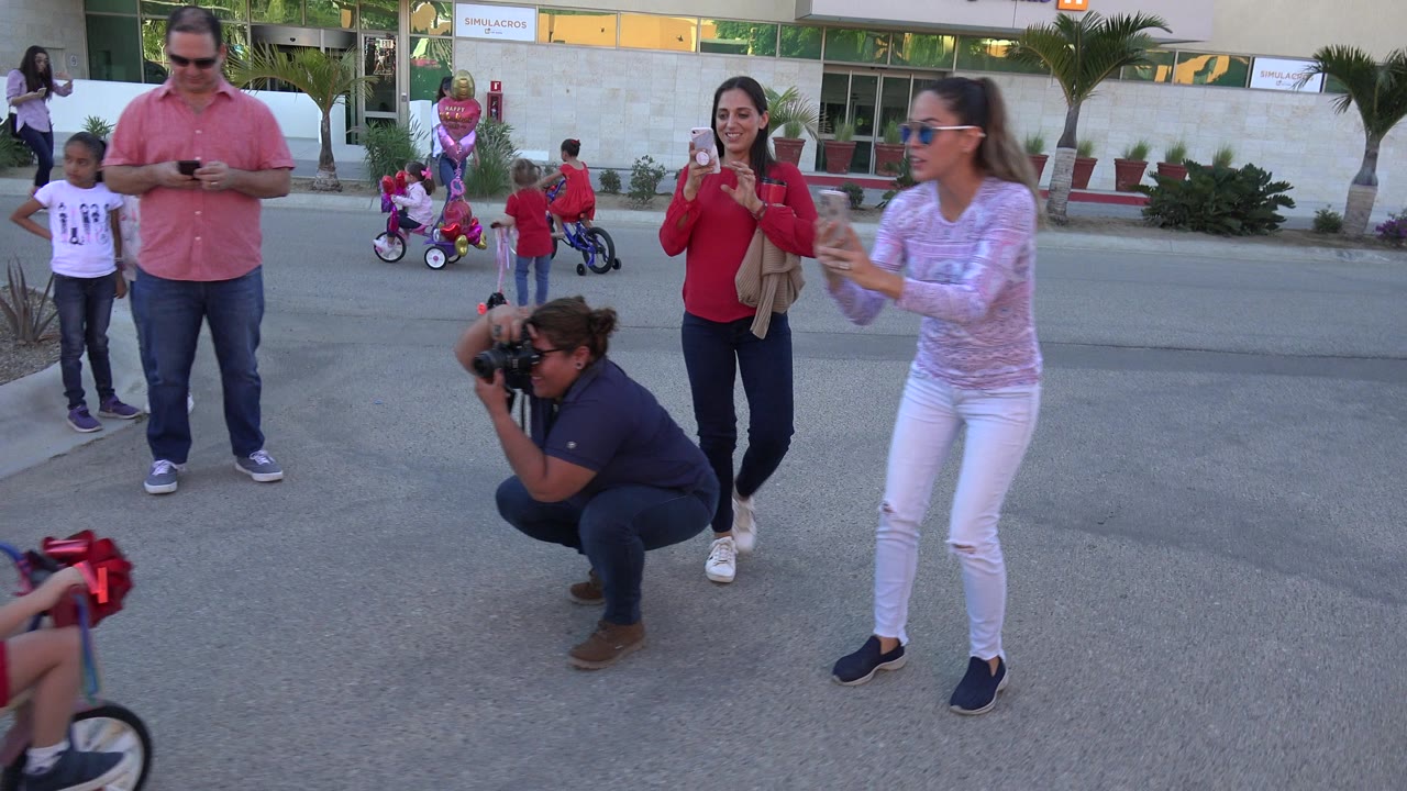 In Los Cabos, Mexico, a man looks down at a phone while women film and children gather around small bikes and pink balloons across the street.