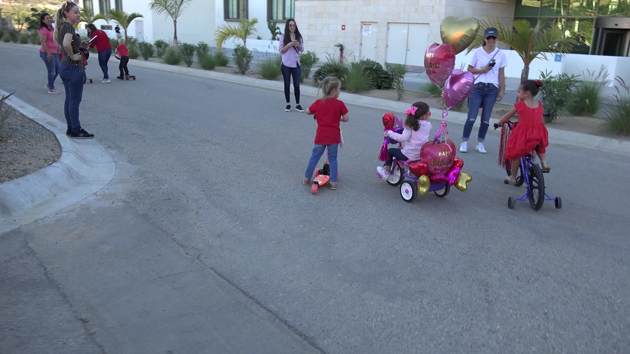 Children ride bikes and a scooter near adults on a paved lane.