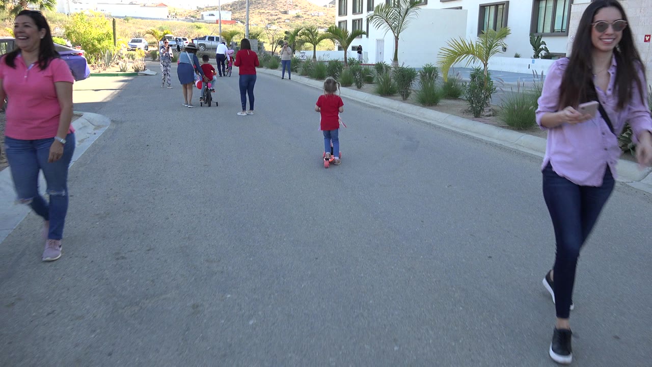 Adults and children on a sunny street with a child riding a red scooter.