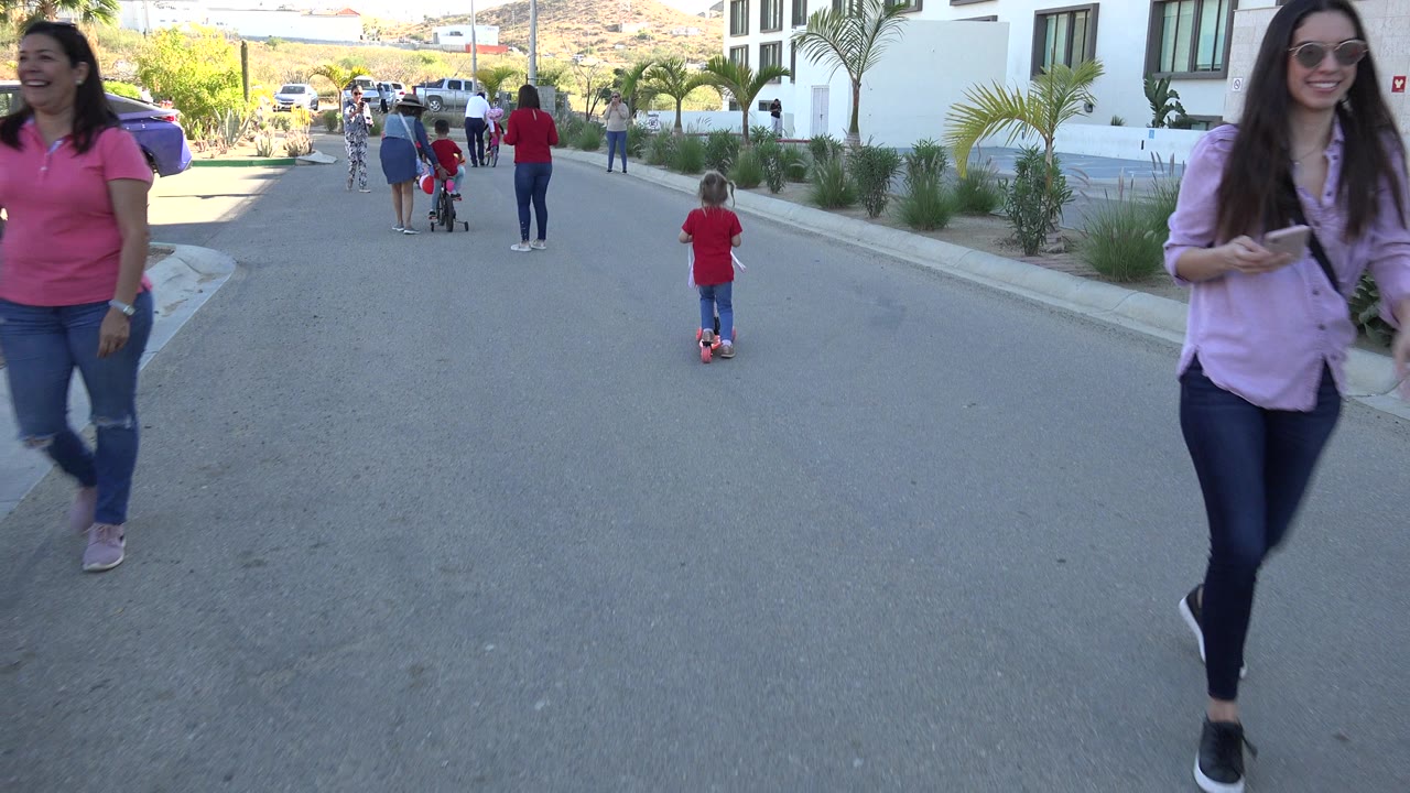 In Los Cabos, two women walk close to the camera while a child on a red scooter continues in the street ahead near parked cars and planted palms.
