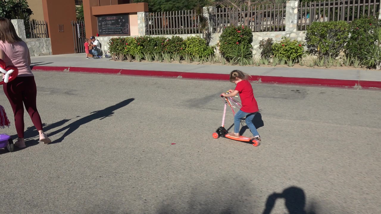 A young girl in a red shirt pushes an orange scooter across a sunlit street in Los Cabos, Mexico, with a woman partly visible at the left edge and a fenced building in the background.