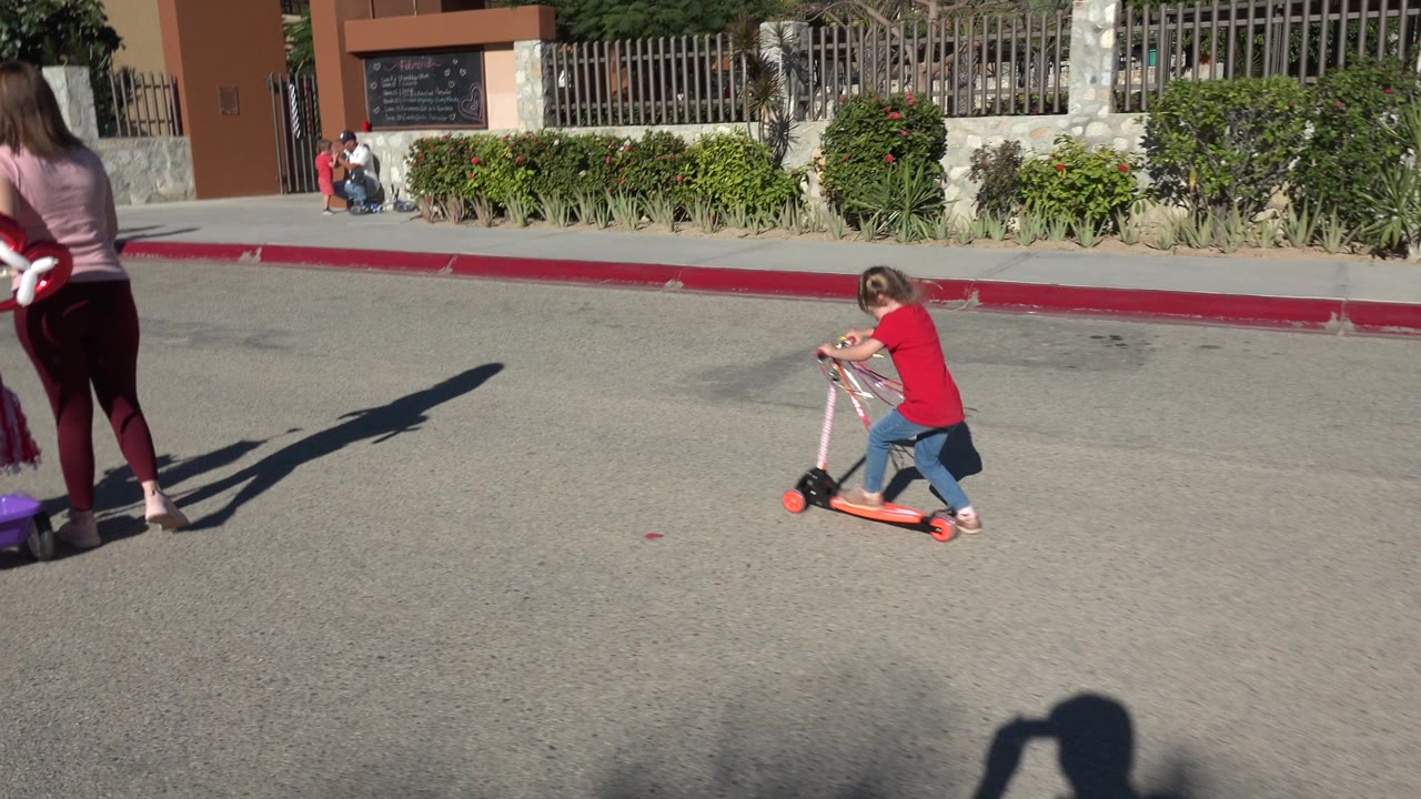 The child continues pushing the orange scooter across open pavement in Los Cabos, Mexico, with a red curb, shrubs, a fence, and distant pedestrians behind her.