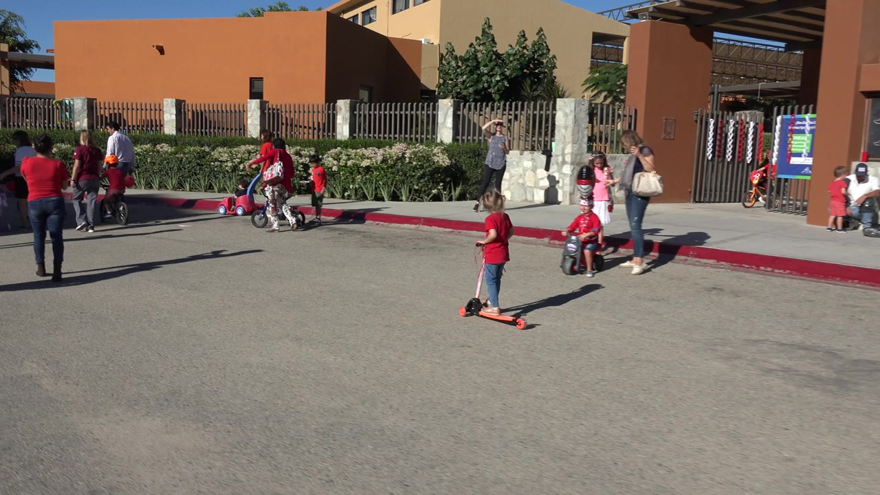 Children and adults on a sunny street while a girl rides a pale pink scooter.