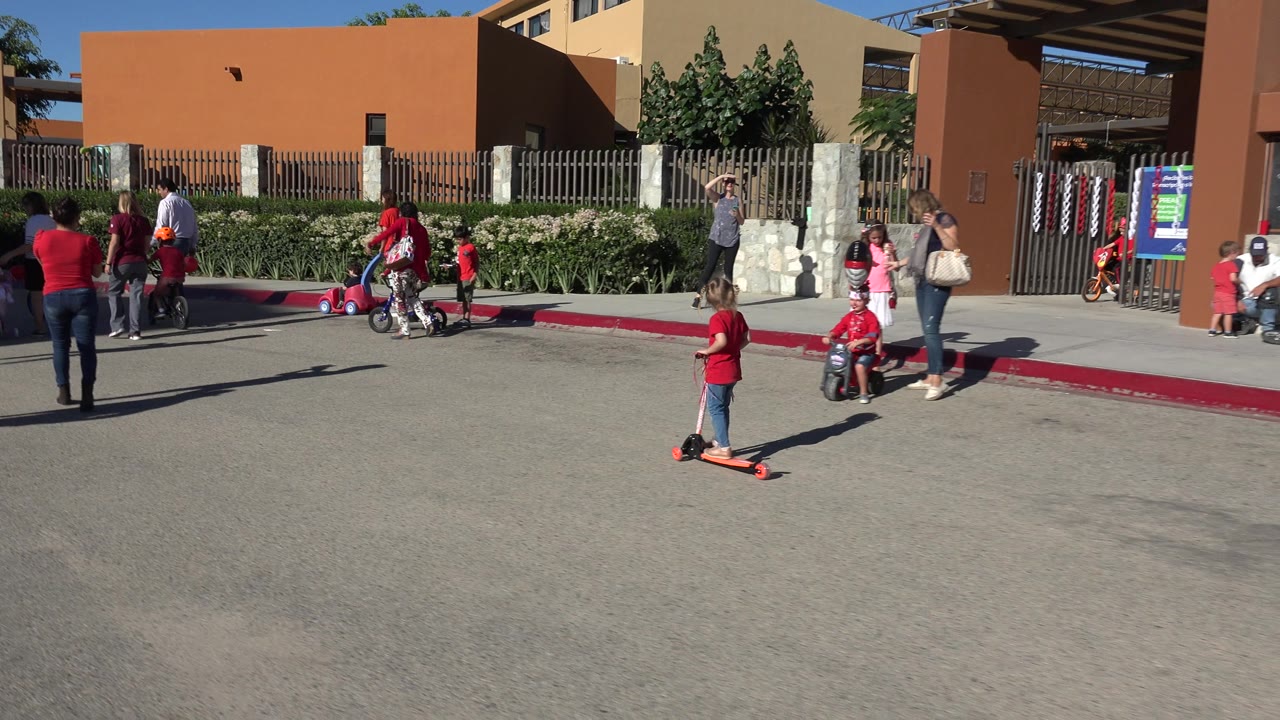 In Los Cabos, Mexico, the girl on the pale pink scooter moves farther across the open road as children on ride-on toys and adults gather near the curbside fence.