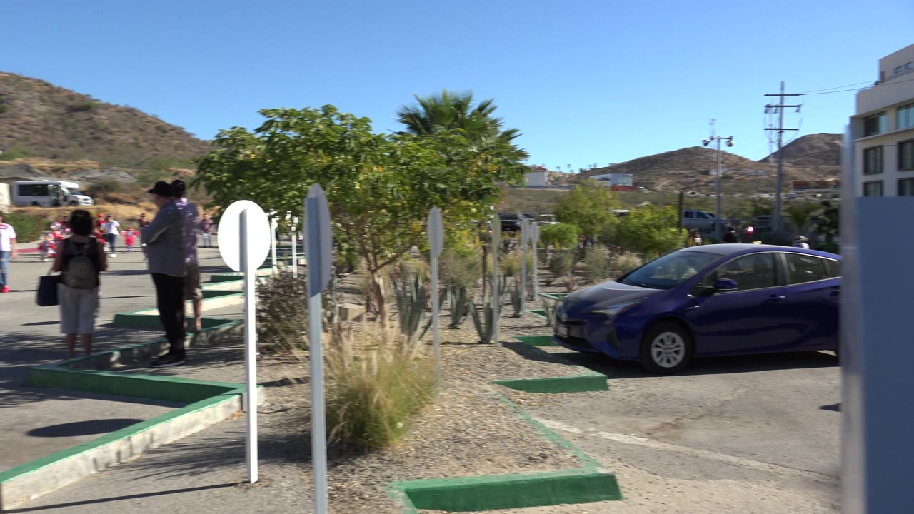 Adults and children stand near a road beside a parked blue car and desert planting.