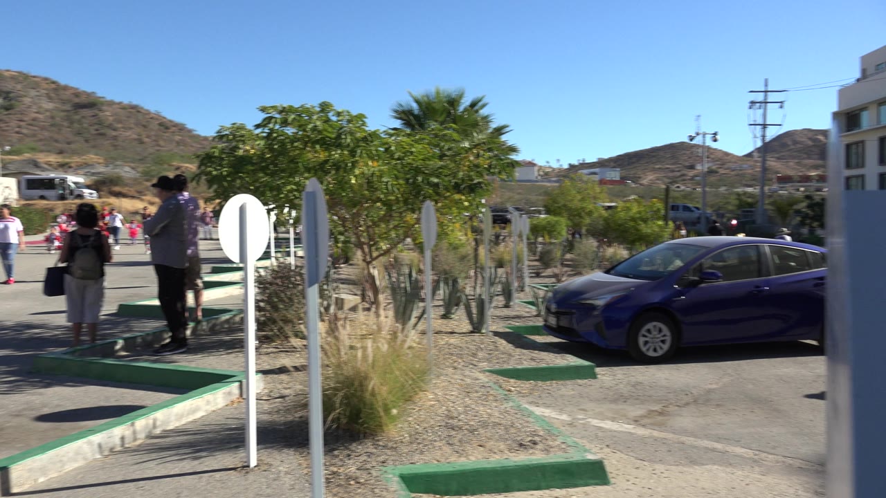 The road scene in Los Cabos, Mexico shows adults and children near a curb, a parked blue car, white signs, desert shrubs, and hillside homes in bright sun.