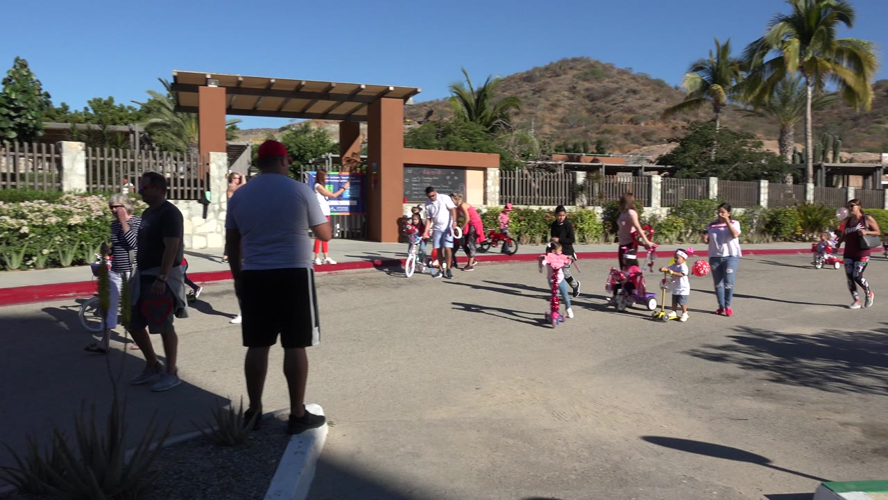 Wide Los Cabos frame showing parents and children near a gated entrance, with scooters, bicycles, hedges, palms, and dry hills behind them.