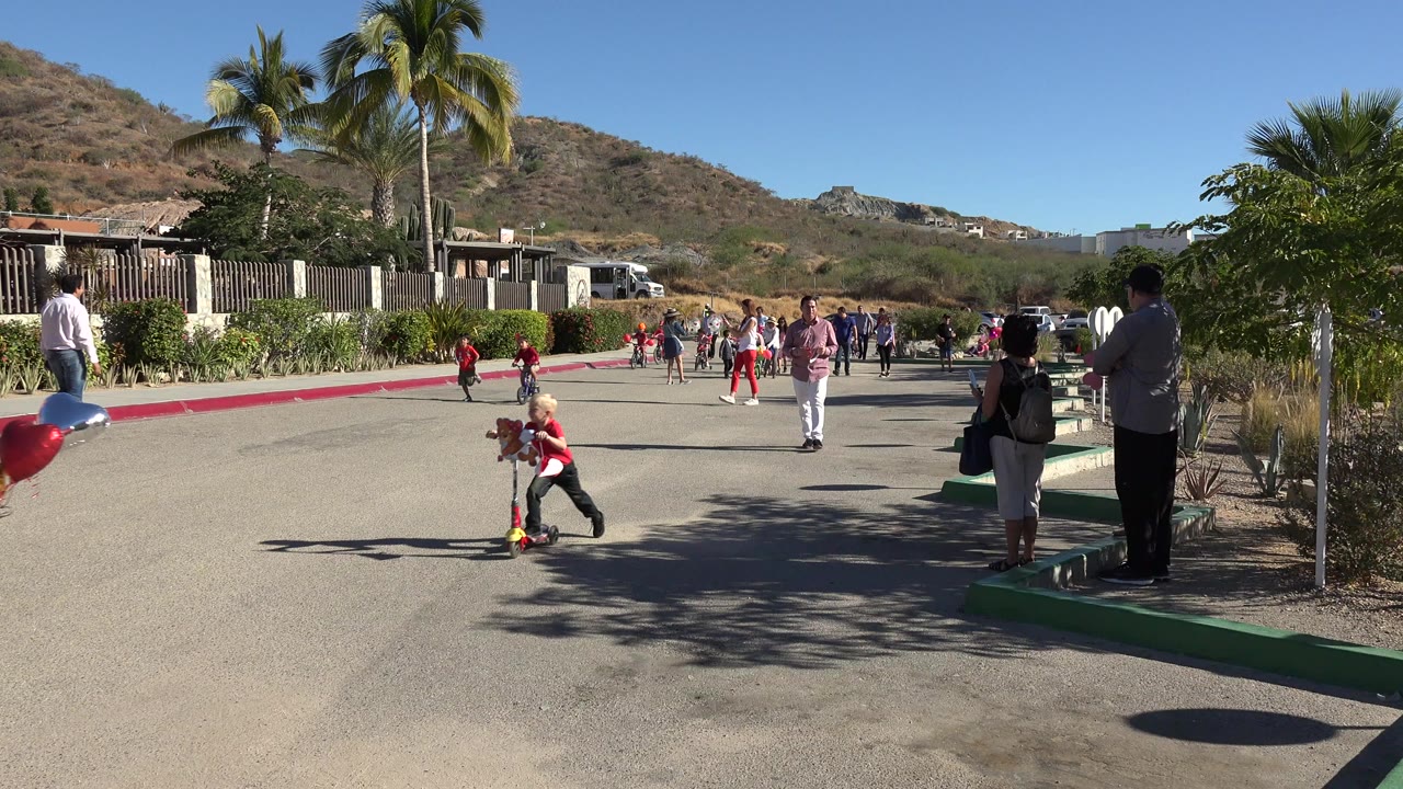 Children ride scooters on a paved road with adults watching nearby.