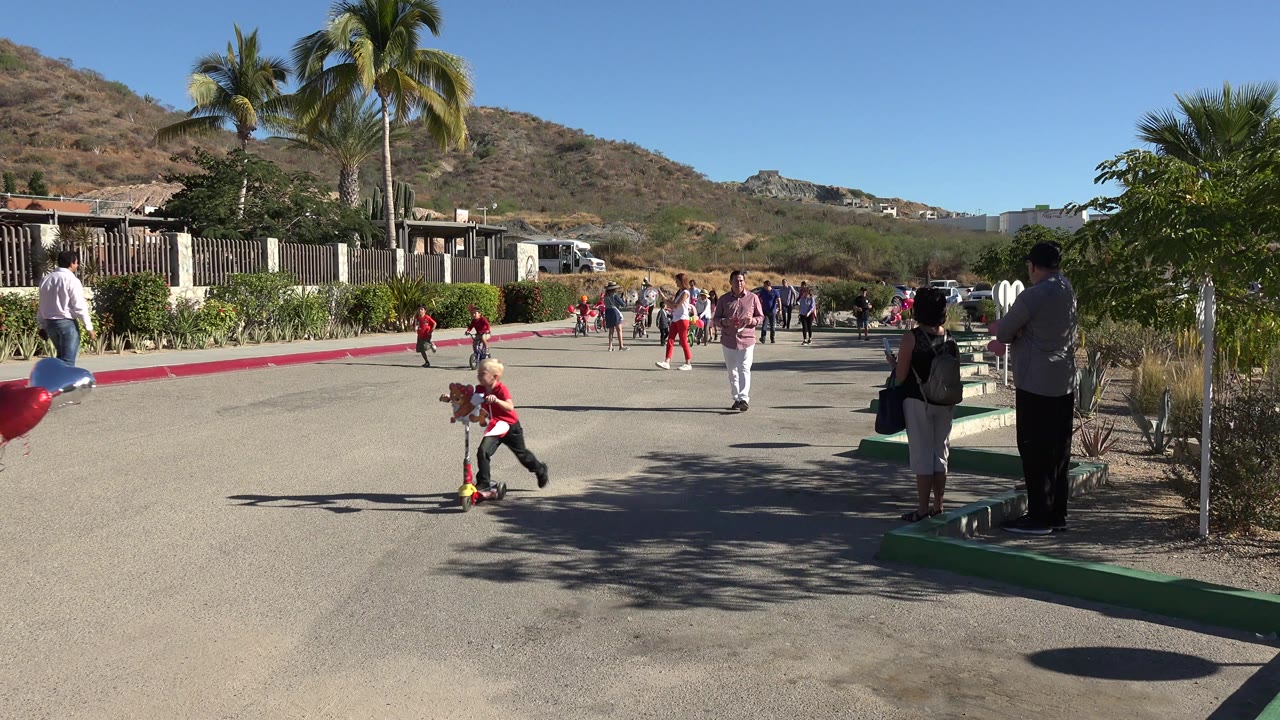 A blond child in a red shirt rides a red scooter toward the right side of the road in Los Cabos, Mexico, while other children and adults remain farther back.