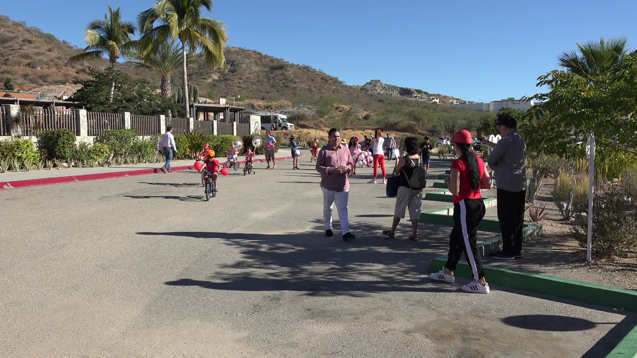 A child wearing an orange helmet rides a small bicycle toward the camera in Los Cabos, Mexico, with red balloons on the handlebars and adults gathered by the curb.