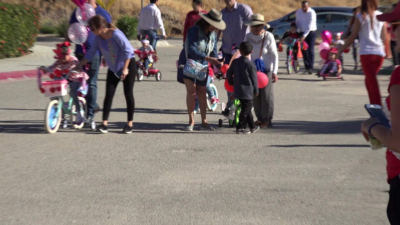 In Los Cabos, Mexico, adults gather around children on small bicycles while a cluster of pink balloons stands near the left side of the road.