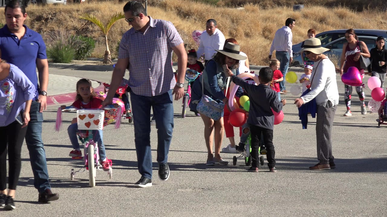 Children with decorated bicycles and balloons on a sunlit road with adults nearby.