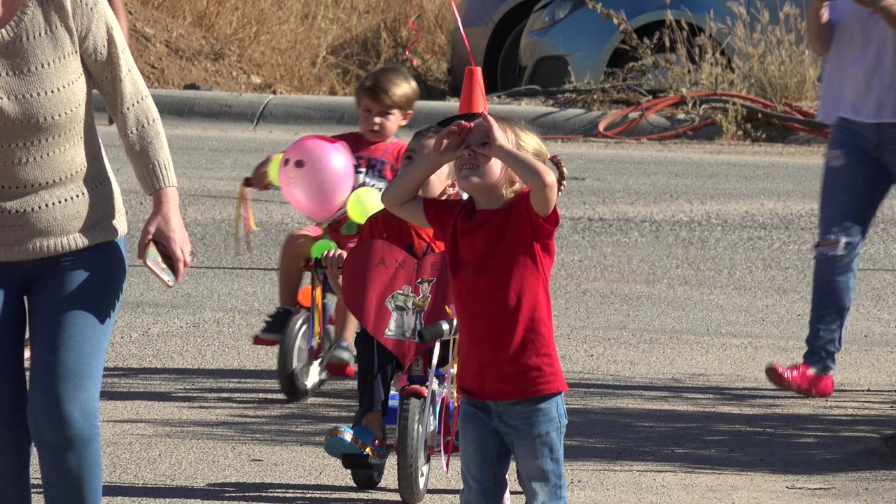 Children ride decorated small bicycles in Los Cabos, Mexico, with an adult in jeans partly foregrounded and dry roadside grass behind them.