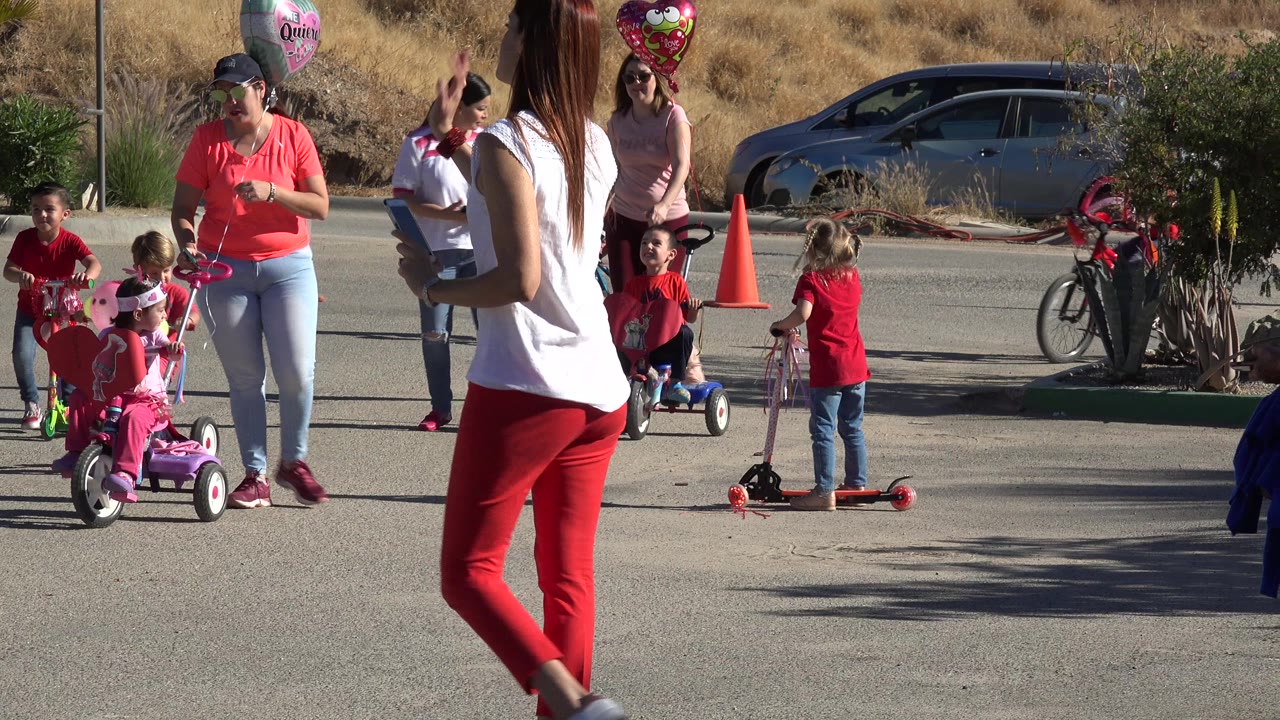 Children and adults on a sunny street with bikes, scooters, balloons, and cones.