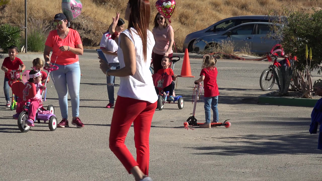 Adults in red and white stand near children on small ride-on toys in Los Cabos, with a silver parked car, cones, balloons, shrubs, and dry grass behind them.