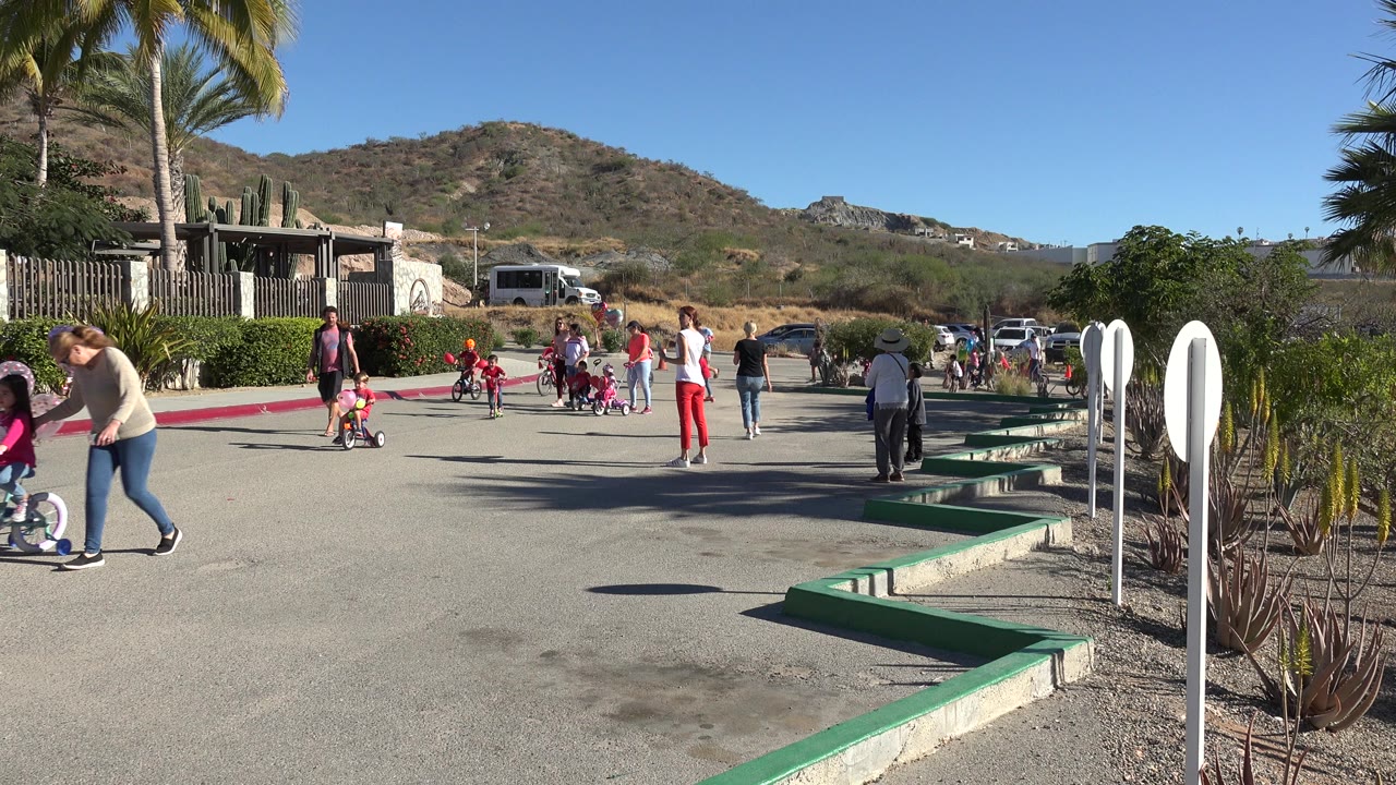 One second in, the Los Cabos street scene shows children riding or standing near small bikes while adults walk nearby, with palms, parked vehicles, and hillside terrain behind them.