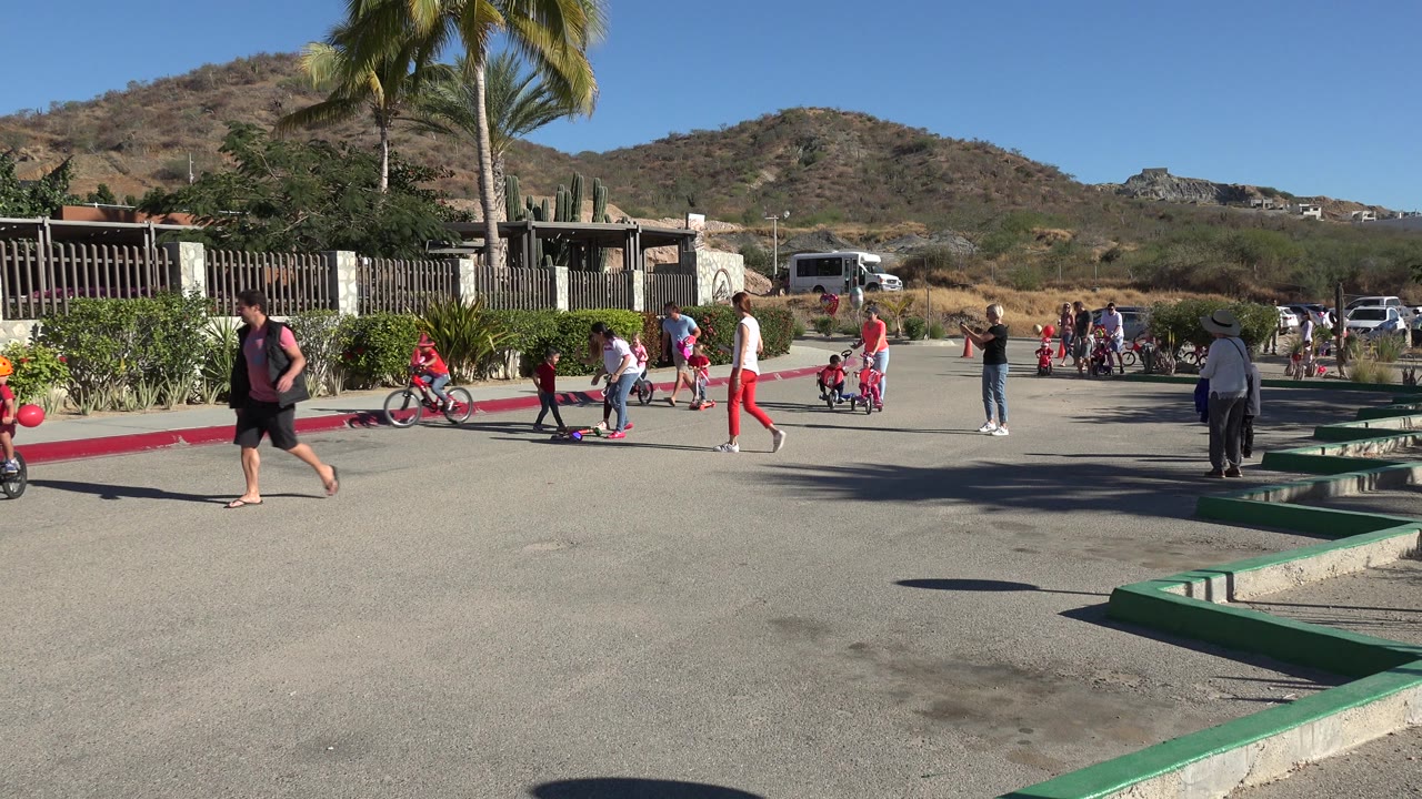 Families and children gather on a paved street near palms and dry hills.