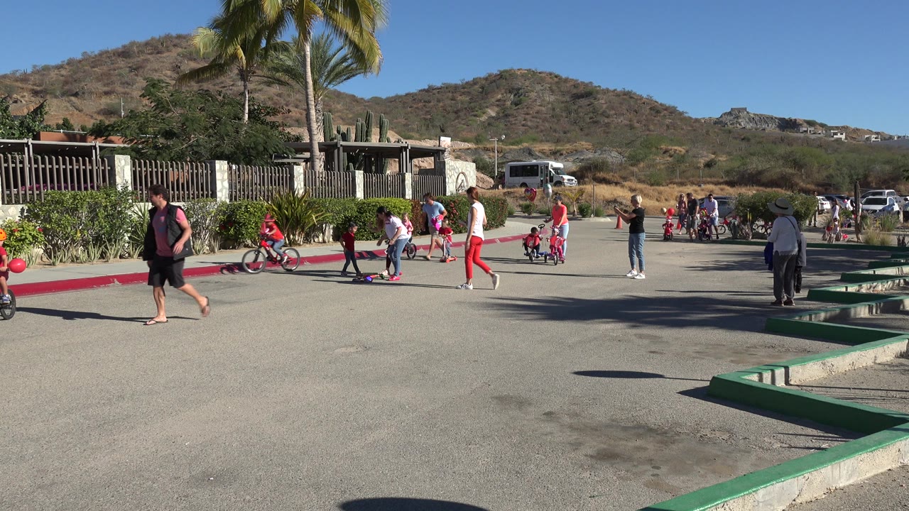 Los Cabos neighborhood street with a child on a red bicycle, other children on scooters, adults watching, a red curb, landscaping, and hills behind them.