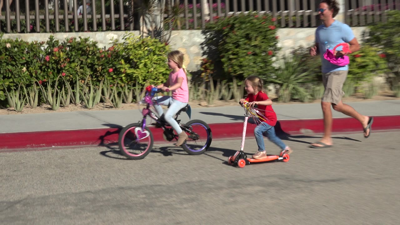 Children ride a bicycle and scooter beside a red curb.