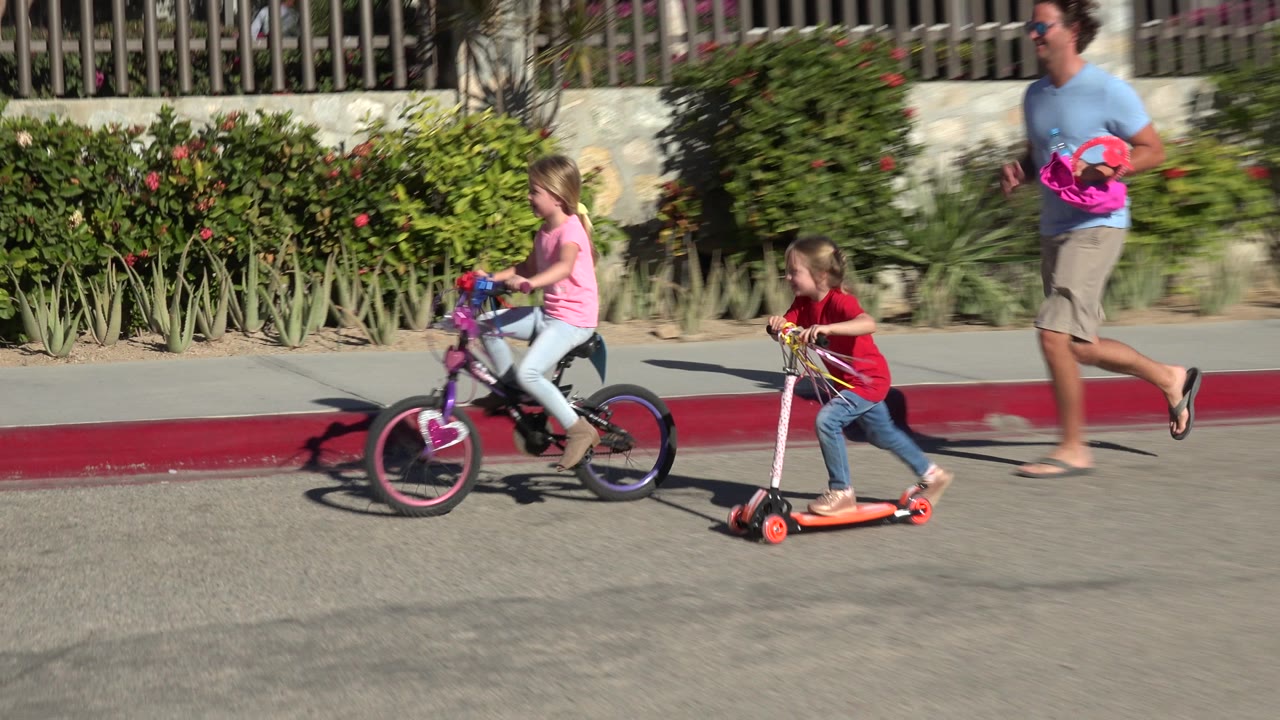 The Los Cabos street scene continues as the bicycling girl leads and the scooter rider follows, with a man carrying a pink item in the background.