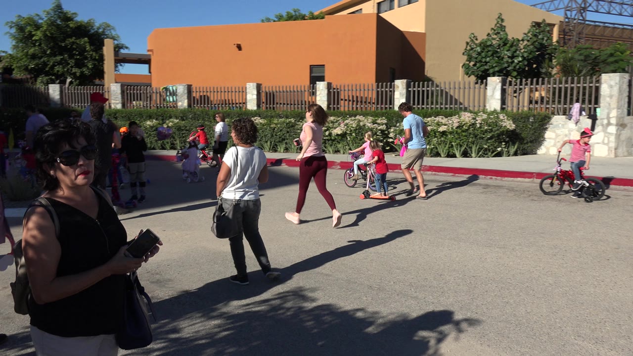 Parents and children move through a sunny paved area near an orange building.