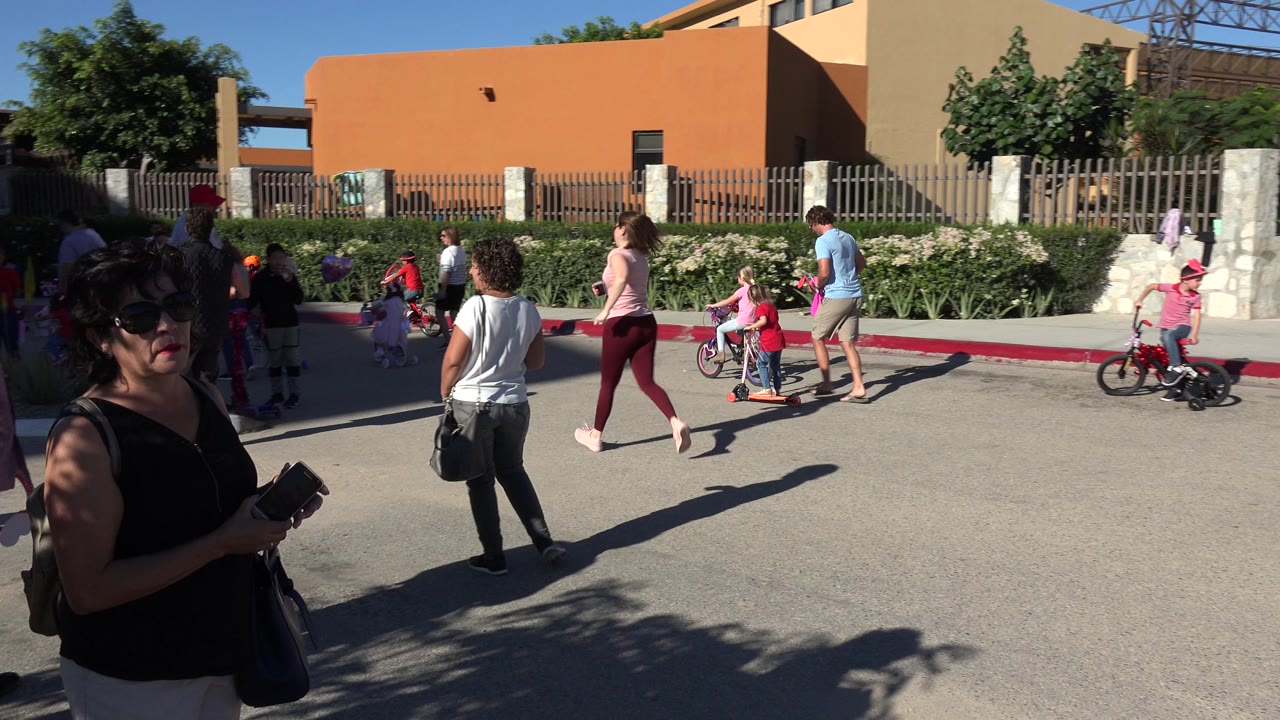A woman wearing sunglasses and holding a phone is close to the camera in Los Cabos, Mexico, while adults and children continue across the paved area behind her.