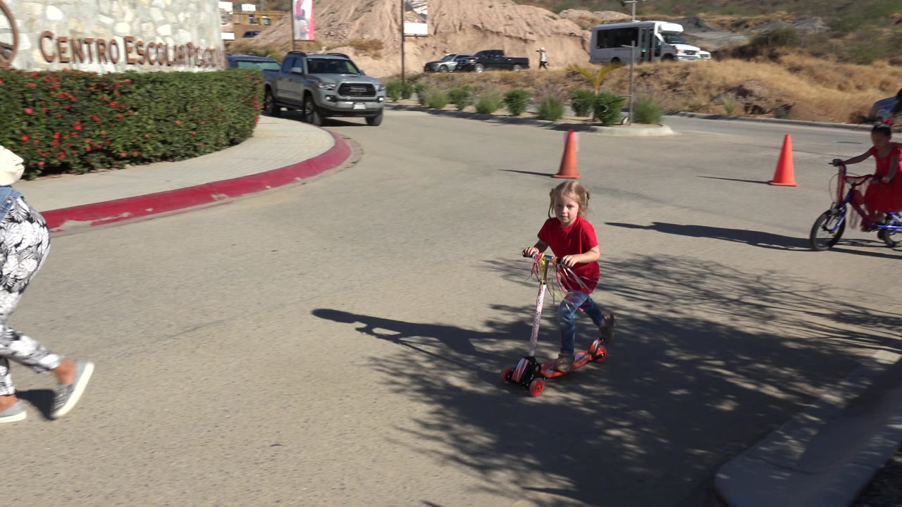 The scooter child moves through the center of the frame on a sunlit Los Cabos street, with orange traffic cones, hedges, parked cars, and a child on a bike in the background.