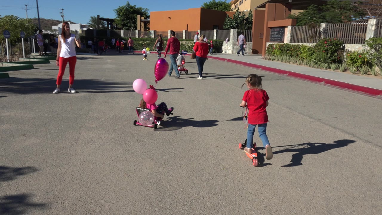 A child in a red shirt rides a scooter near a cart with pink and heart-shaped balloons as families walk ahead on a bright Los Cabos street.